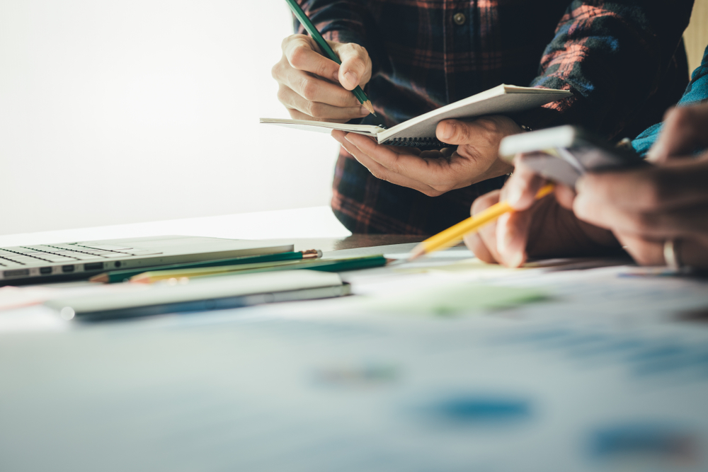 2 pairs of hands holding a notebook and Calculator over a desk with office supplies on it